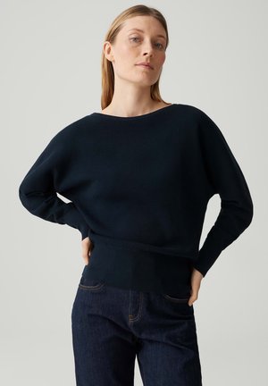 Donna con capelli chiari che indossa un maglione a maniche lunghe blu navy scuro e jeans blu scuro, con le mani appoggiate sui fianchi su uno sfondo semplice.