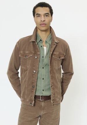 Moški, ki nosi rjavo denim jakno in hlače ter zeleno srajco s sponkami, stoji z rokami za hrbtom na belem ozadju.