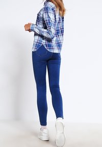 Modrá a bílá kostkovaná košile s dlouhými rukávy a zaobleným spodním okrajem. Kombinována s přiléhavými tmavě modrými džíny a bílými teniskami.