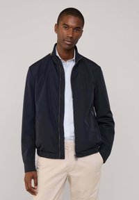 Chaqueta de cremallera ligera en color navy con cuello alto, puños y cintura acanalados, que presenta bolsillos laterales y un ligero brillo. Lleva puesta sobre una camisa de rayas.