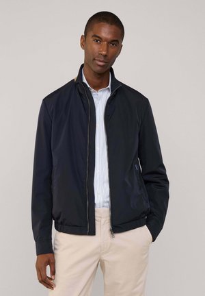 Veste zippée légère navy avec un col montant, des poignets et une taille côtelés, dotée de poches latérales et d'un léger brillant. Portée par-dessus une chemise rayée.