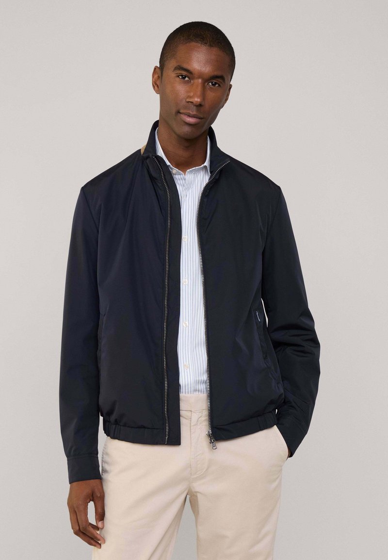 Chaqueta de cremallera ligera en color navy con cuello alto, puños y cintura acanalados, que presenta bolsillos laterales y un ligero brillo. Lleva puesta sobre una camisa de rayas.