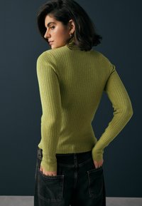 Rippstrickpullover in Grün mit hohem Kragen und langen Ärmeln, kombiniert mit dunkler Jeans. Glatte Textur, enganliegendes Design.