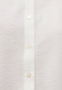 White button-up shirt, lightweight textured fabric, featuring a vertical row of seven transparent buttons along the front.