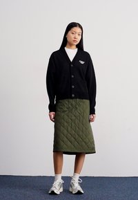 Cardigã preto tricotado com decote em V, fecho de botão e logótipo. Combinado com uma saia verde acolchoada e sapatilhas brancas com detalhes cinzas.