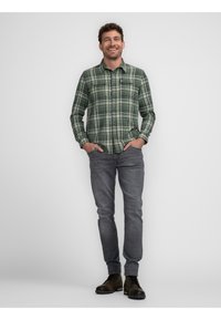 Camisa a cuadros verde y negra con botones, dos bolsillos en el pecho, jeans gris y botas oscuras, con un modelo masculino de pie frente a un fondo plano.