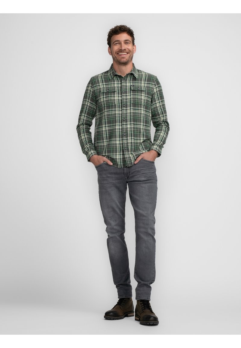Camisa a cuadros verde y negra con botones, dos bolsillos en el pecho, jeans gris y botas oscuras, con un modelo masculino de pie frente a un fondo plano.