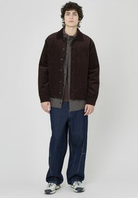 Veste en velours côtelé marron avec deux poches poitrine, portée par-dessus une chemise à carreaux. Pantalons en denim foncé avec couture visible et chaussures de sport.
