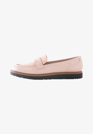 Persoon met lichtblauwe opgerolde jeans en roze leren loafers staand op een grijze vloer.