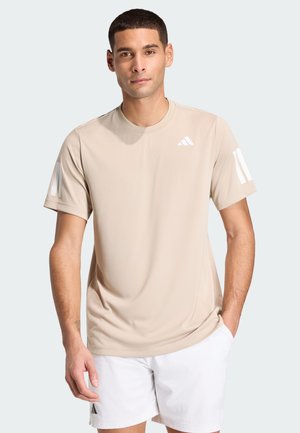 Homme debout, les mains dans les poches, portant un t-shirt beige Adidas et un short blanc sur fond clair.