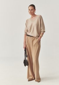 Beige, V-nyakú pulóver laza ujjal; világosbarna, bő szárú nadrág zsebekkel; fekete öv; kicsi fekete kézitáska; hegyes barna cipők.