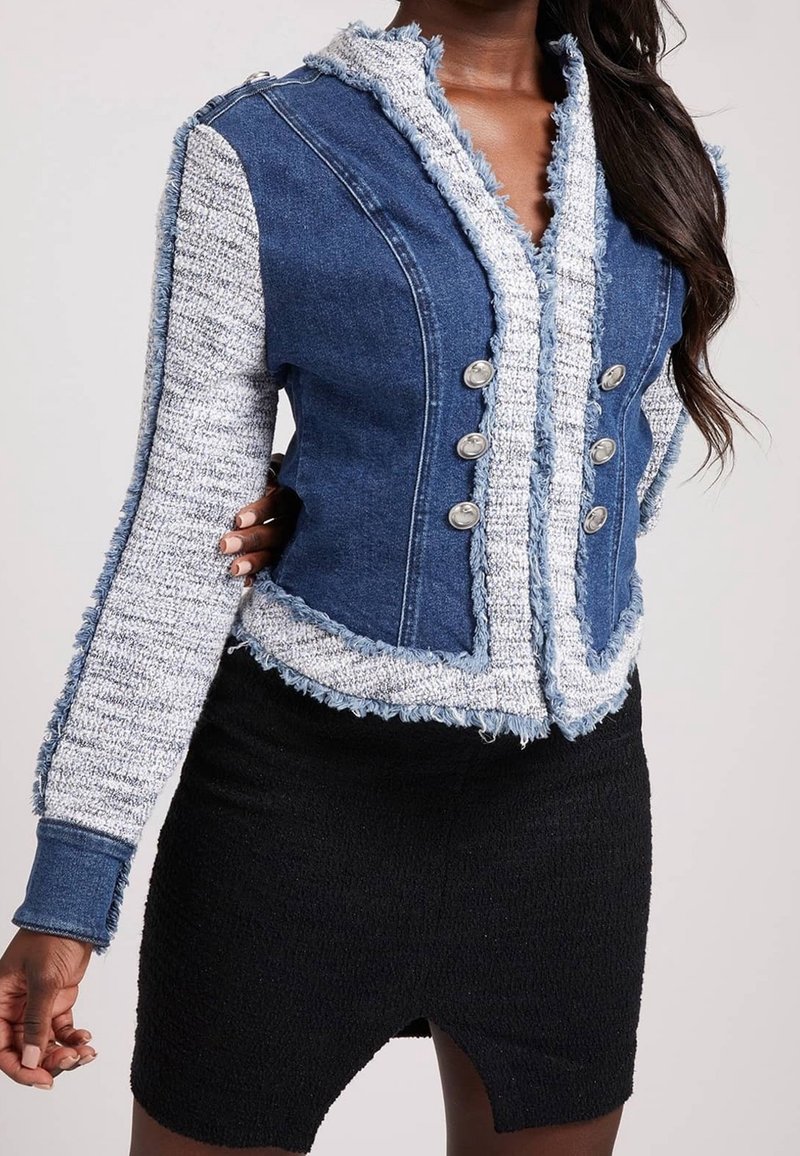 Femme portant une veste en jean ajustée avec des bords texturés et effilochés et des boutons argentés sur une jupe noire texturée avec une fente à l’avant.