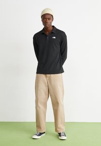 Svart langermet polo skjorte med krage og subtilt logo; kombinert med beige bukser og svart-hvite joggesko på et grønt gulv.