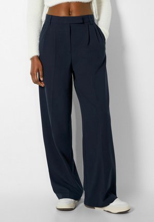 Navyblauwe wijd uitlopende broek met hoge taille en plooien aan de voorkant, gedragen met witte sneakers en een kort, wit, pluizig vest, handen in de zakken.