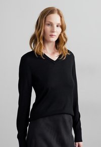 Črni pulover z v-neck iz mehkega pletenega materiala, z dolgimi rokavi in sproščenim krojem. Obut z črno krilom na nevtralni ozadju.