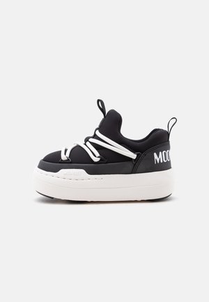 Zapatilla de plataforma en blanco y negro con suela gruesa, cordones blancos cruzados y lengüetas para tirar en la lengüeta y el talón.