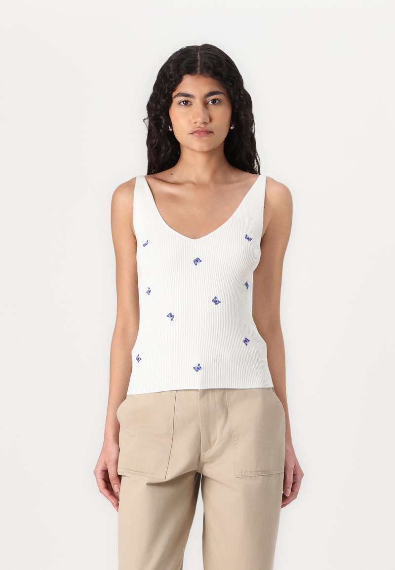 Ribbelde witte tanktop met een diepe V-hals en blauwe vlinderborduring. Gecombineerd met beige hoge taille broek met een relaxte pasvorm.