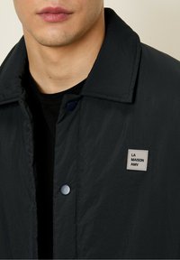 Black lightweight jacket with a smooth finish, featuring a collar, snap buttons, and a small, square brand tag on the chest.