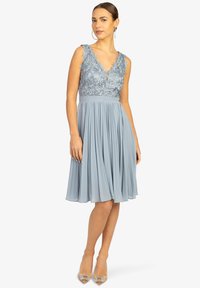 Robe bleu clair avec un corsage ajusté en dentelle, décolleté en V profond et jupe évasée plissée. Finition avec une délicate broderie florale.