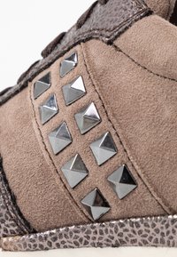 Bruna mocka sneakers med metalldekor, en texturerad ovandel med reptilmönster och en kontrasterande glänsande kant längs sulan.