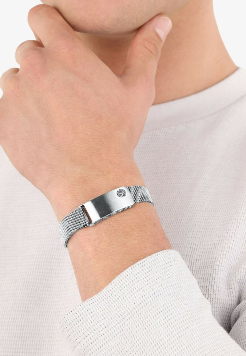 Pulsera de malla de plata con un cierre de metal pulido, que presenta un pequeño emblema. El diseño es elegante con un acabado texturizado en la banda.