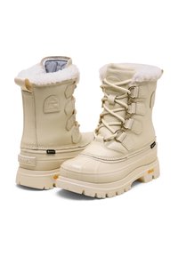 Beige talvikengät paksuilla kumipohjilla, nauhakiinnityksellä, lampaankarvakauluksilla ja Gore-Tex-tunnuksilla vedenpitävyyden varmistamiseksi.