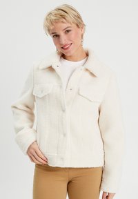 Veste en polaire blanche avec une fermeture à boutons sur le devant, des poches poitrine et un col. Elle présente une texture douce et une coupe décontractée.