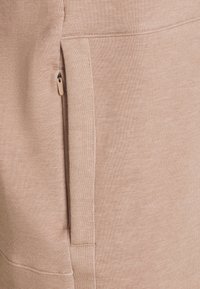 Tejido de sudadera beige claro con cremallera lateral y textura suave. Los detalles de las costuras añaden una dimensión sutil.
