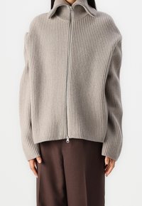 Maglione in maglia con zip di colore grigio chiaro, texture a coste, colletto alto e spalle scese. Abbinato a pantaloni larghi marroni.