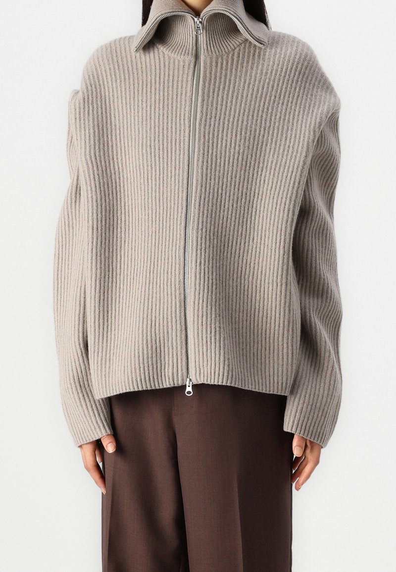 Maglione in maglia con zip di colore grigio chiaro, texture a coste, colletto alto e spalle scese. Abbinato a pantaloni larghi marroni.