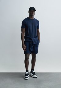 T-shirt à manches courtes bleu marine foncé avec un logo subtil, associé à un short assorti et un sweat-shirt noué à la taille. Des baskets noires complètent la tenue.