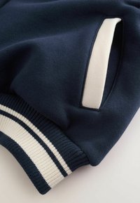 Marineblå hættetrøje med detaljer, der viser et glat stof med en cremefarvet detalje nær lommen og en ribbet kant med to hvide striber.