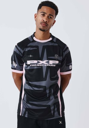 Uomo con capelli corti e ricci e orecchini in piedi, rivolto in avanti, indossando una maglietta nera e grigia Project X Paris con bordi rosa.