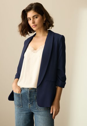 Mujer con cabello corto y oscuro que lleva una blazer azul marino sobre una camiseta blanca con encaje y vaqueros azules, de pie frente a un fondo neutro.