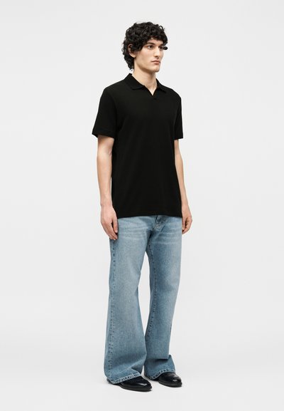 Jeune homme debout portant un polo noir à manches courtes, un jean large bleu clair et des chaussures noires sur un fond uni.