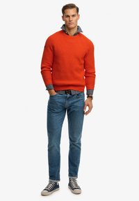 Suéter de punto naranja con textura acanalada, combinado con una camisa de cuadros azul y blanca, jeans ajustados de mezclilla y zapatillas deportivas azul marino.