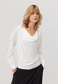 Kobieta z blond włosami, ubrana w białą drapowaną bluzkę, czarną spódnicę, złotą bransoletkę i złote kwiatowe kolczyki, pozująca z rękami w kieszeniach.