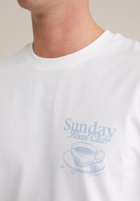Balts kokvilnas T-krekls ar gaiši zilu grafiku, uz kuras attēlots kafijas krūze ar tekstu "Sunday Roast Club" virs tās, īsām piedurknēm un apkakli.