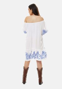 White off-the-shoulder dress with blue floral embroidery on sleeves and hem, features a tiered ruffle at the bottom and a relaxed fit. Paired with brown cowboy boots.