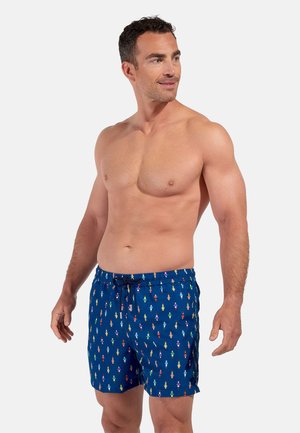 Shirtless man wearing blue swim shorts with colorful person icon pattern, standing and looking to his right.