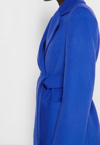 Abrigo de lana azul con cuello de solapa, cintura ceñida con cinturón y bolsillos laterales. Textura suave con un ajuste entallado y mangas largas.