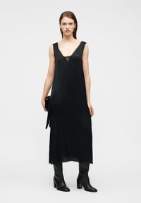 Γυναίκα φορόντας μαύρο αμάνικο φόρεμα μήκους μέχρι τη μέση της γάμπας με δαντέλα, μαύρες ψηλές μπότες μέχρι τη μέση της γάμπας και κρατώντας μαύρο τσαντάκι με φουντίτσες.