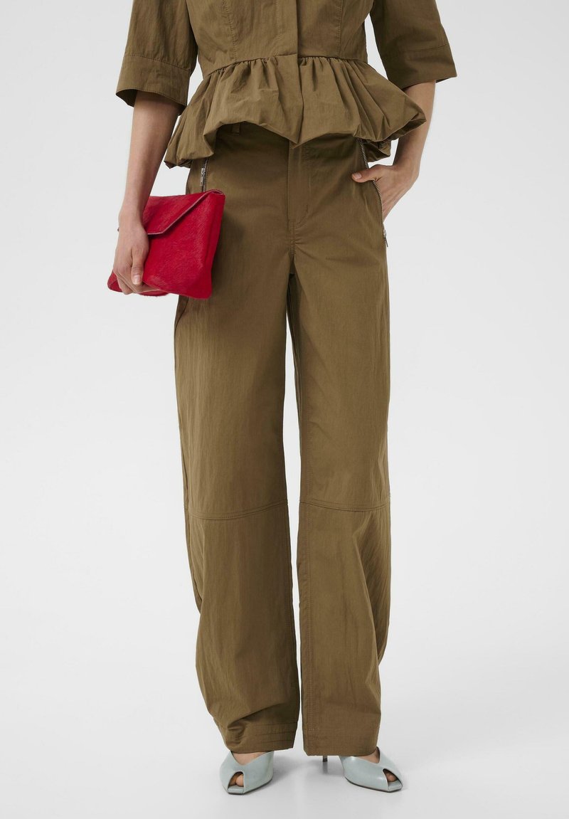 Vrouw draagt bruine wijde broekspijpen en een peplumtop, houdt een rode clutch vast, staat op lichtblauwe open teen hakken tegen een effen achtergrond.