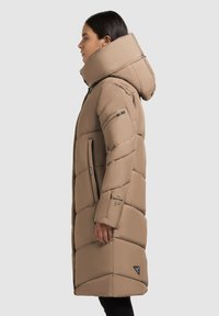 Kvinde iført en lang, beige polstret vinterjakke med høj krave og hætte, som står i profil mod en ensfarvet lys baggrund.