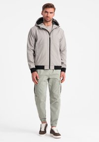 Giacca bomber grigia chiara con cappuccio, cerniera nera e polsini a coste; abbinata a pantaloni cargo verde chiaro e sneaker marroni con lacci.