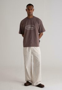 Brown cotton T-shirt with a white logo, paired with white and beige striped wide-leg pants, and dark brown slip-on shoes.