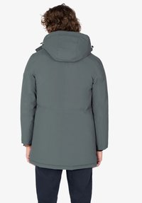 Persona con cabello rizado que lleva una chaqueta de invierno con capucha gris y pantalones oscuros, de espaldas contra un fondo blanco liso.