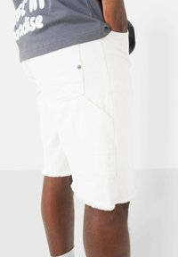 Shorts in denim bianco con orlo sfrangiato, taglio dritto e tasche laterali. Indossati con una maglietta grigia con testo bianco.