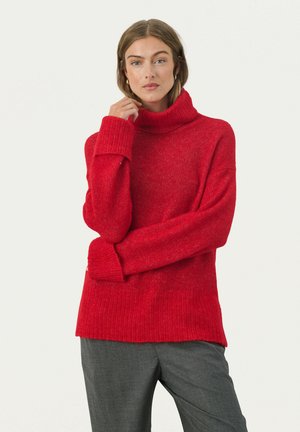 Roter Strickpullover mit hohem Kragen, gerippter Textur und oversized Passform. Lange Ärmel mit breiten Bündchen. Graue Hose ergänzt das Outfit.