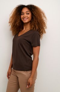 Camiseta de algodón marrón con escote en V y mangas cortas, combinada con pantalones beige claros. El modelo está de lado, mostrando un ajuste relajado.